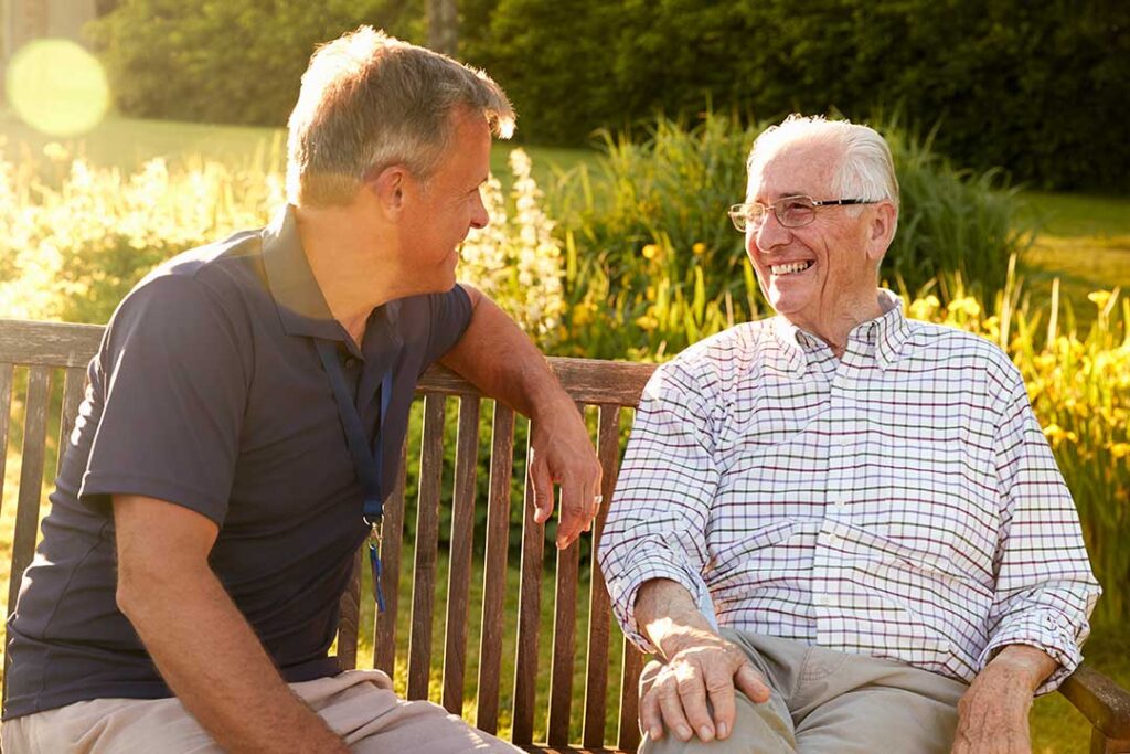 Two older adults sitting in a sunny common area, talking and laughing together over coffee.