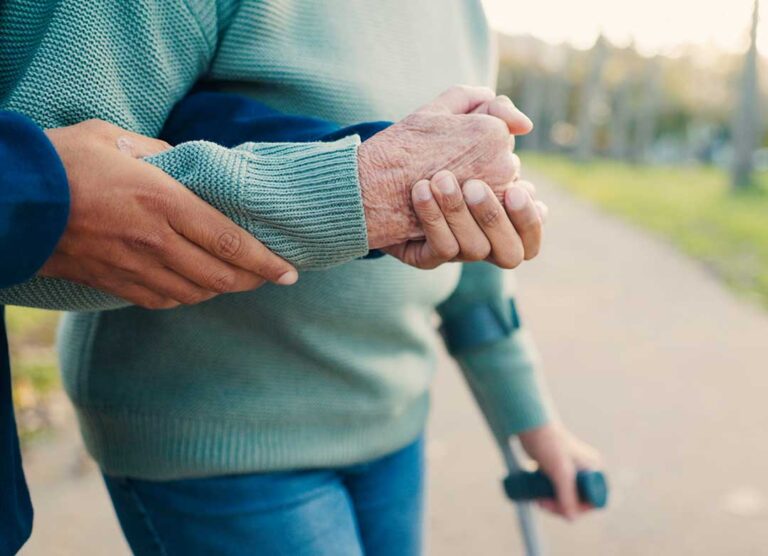 Caregiver helps an older resident walk safely with a walker through a bright common area.